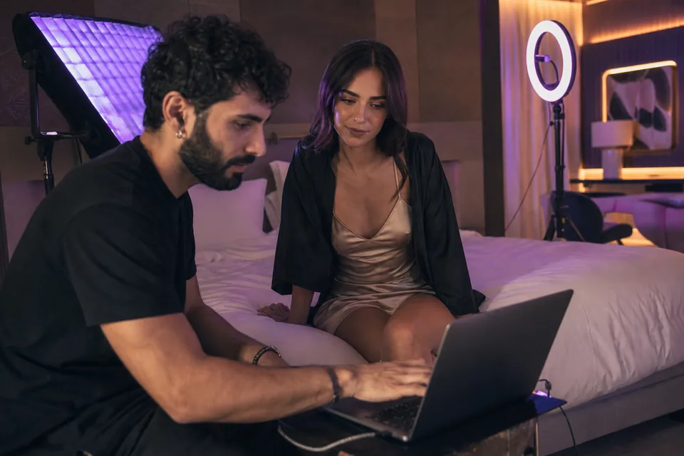 Photographer reviewing shots on a laptop with a woman in a satin dress during a content shoot in a hotel suite